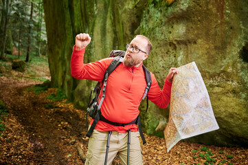 Bearded tourist man studies map while stands in front of large rock in dense forest. Traveler with...