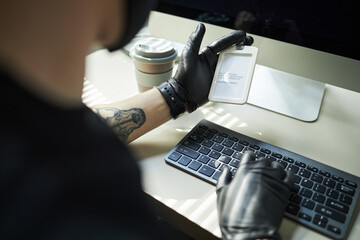 Young male data thief wearing black gloves holding identification badge while typing password on...