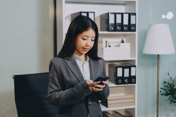Young, smiling Asian businesswoman holding a jacket on her shoulder while using a mobile phone,...