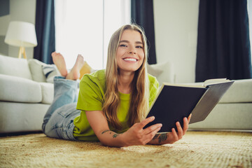 Young woman lying on the floor in a cozy living room enjoying a book, dressed casually in a green...