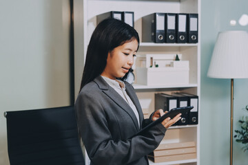 Young, smiling Asian businesswoman holding a jacket on her shoulder while using a mobile phone,...