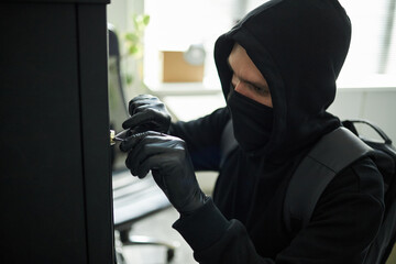 Young Caucasian man wearing black hoodie and mask picking lock on cabinet with gloved hands in...