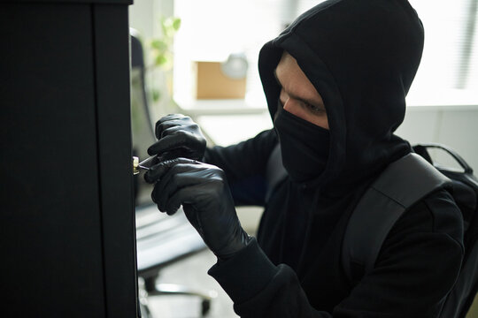 Young Caucasian man wearing black hoodie and mask picking lock on cabinet with gloved hands in office setting, backpack on shoulders, to steal confidential business documents