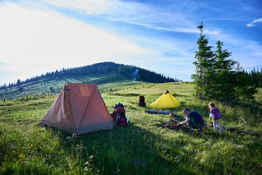 Picturesque mountain campsite on grassy hill with tents. Camper and child tend to campfire, surrounded by backpacks and gear. Rolling hills, evergreens, clear blue sky. Peaceful outdoor adventure.