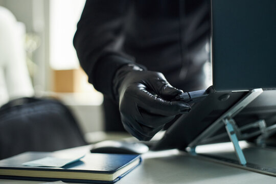 Anonymous man wearing black gloves inserting USB flash drive into laptop computer, performing data transfer, theft, malware or cyber fraud in modern corporate office
