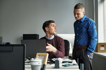 Caucasian young adult man sitting at desk explaining project to team member standing nearby in...