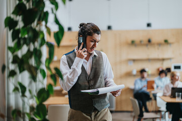 Businessperson reviewing papers while conversing on phone in shared workspace. Collaborative...