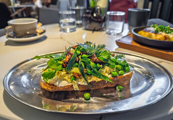 An upscale brunch dish: A piece of rustic sourdough toast topped with egg scramble, sauteed mushrooms, and fresh greens on a marble table in a cafe.