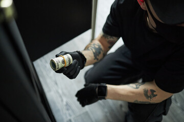 Tattooed young man wearing black gloves kneeling on floor stealing roll of US dollar bills from...