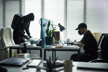 Two young male burglars in dark clothing hacking into corporate networks on laptops in company...
