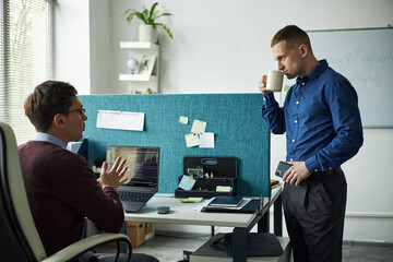 Caucasian young man sitting at desk discussing project with male team colleague standing and drinking from mug in modern office workspace, discussing work project, with laptop and documents visible