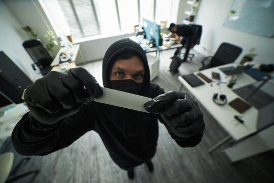 Caucasian young man, wearing black hoodie and gloves, covering security camera with adhesive tape during burglary in company office, while accomplice is searching desk for business documents