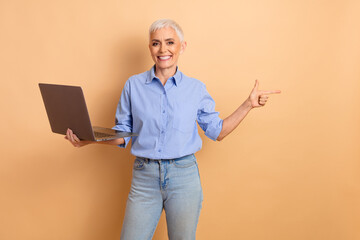Confident senior woman gesturing while holding a laptop in casual attire against a beige background