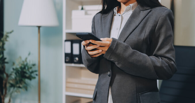 Young, smiling Asian businesswoman holding a jacket on her shoulder while using a mobile phone, standing confidently in a stylish, modern office environment