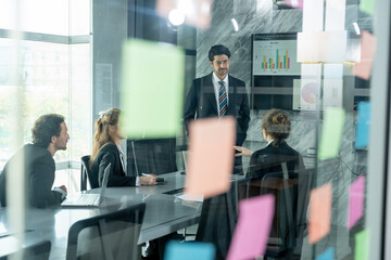 corporate team working together during business meeting inside glass room with multiracial employees brainstorming and listening to male asian presenter in front of digital chart and reports