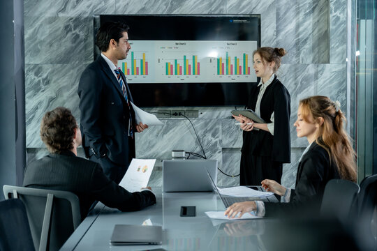 Business professionals engaging in active discussion during team meeting, caucasian woman standing near screen presenting data while female coworker typing notes, multiracial group working together