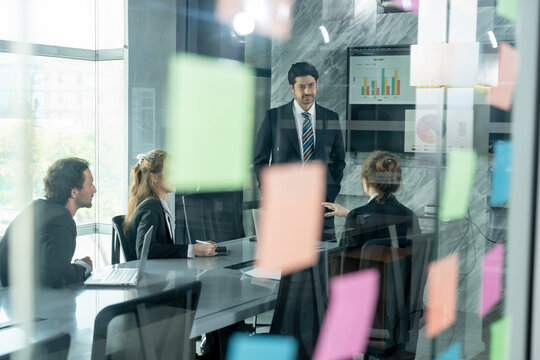 corporate team working together during business meeting inside glass room with multiracial employees brainstorming and listening to male asian presenter in front of digital chart and reports - Powered by Adobe