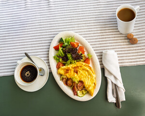 Hearty morning meal of fluffy egg omelette draped over sauteed vegetables and bacon paired with fresh lettuce and tomato salad