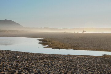 Surfers and beachgoers walk along the shoreline at sunset, immersed in misty golden light. The warm...