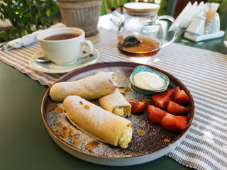 A delicious gourmet breakfast with sweet cheese-filled crepes (blini), fresh strawberries, almond flakes, and sour cream on a plate on a sunlit cafe table.Morning breakfast scene
