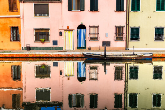 View of colorful buildings mirrored in the tranquil canal waters, with a small boat gently floating creating a vibrant, picturesque scene, Comacchio, Emilia-Romagna, Italy.