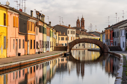 View of vibrant, colorful houses line a tranquil canal beneath an arched bridge, reflections shimmering in the water, Comacchio, Emilia-Romagna, Italy.