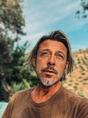 Tanned man with blond half long hair and grey stubble beard in brown t-shirt enjoys sunny summer weather in garden in Spain.