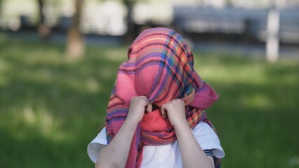 A child, facing away from the camera, enjoys a playful moment by covering their head with a brightly colored scarf, surrounded by a lively park atmosphere.