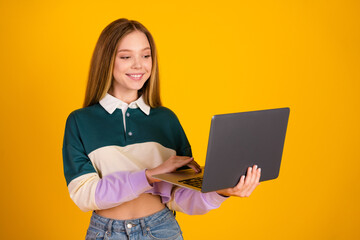 Cheerful young woman in casual clothing holding a laptop against a yellow background, showcasing...
