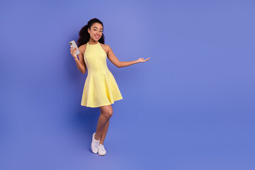 Cheerful young woman with smartphone in yellow dress against purple background showcasing modern...
