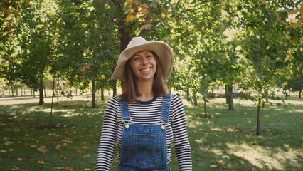 A woman wearing a striped long-sleeve shirt and denim overalls smiles in a vibrant park filled with green trees and autumn-colored leaves on a sunny day.
