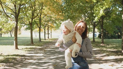Smiling mother strolls through a park, carrying a baby in a cozy outfit while enjoying the warm sunlight filtering through vibrant autumn leaves.