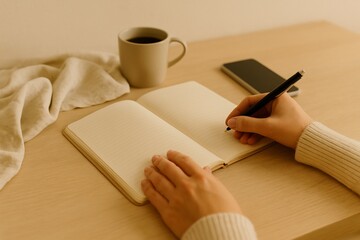 Top-down view of a woman’s hands writing in a notebook. Cozy desk setup with coffee cup and smartphone. Warm natural tones, minimal home interior, no face visible.