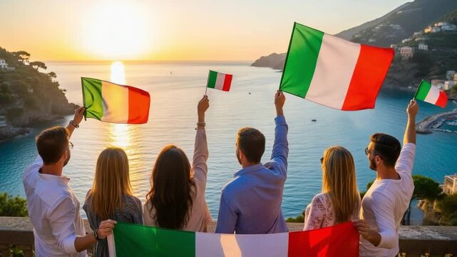 Group of six young friends holding Italian flags on a terrace overlooking a beautiful coastal town at sunset.