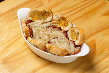 Delicious portion of sliced ​​pepperoni fried with onion and slices of bread (Por&ccedil;&atilde;o de Calabresa Frita) served in a white ceramic bowl on a wooden table.