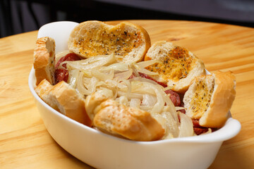 Delicious portion of sliced ​​pepperoni fried with onion and slices of bread (Porção de Calabresa Frita) served in a white ceramic bowl on a wooden table.