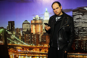 A guy in a black leather jacket with a large hood and black pants with a phone in his hands against...