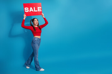 Young woman in stylish casual outfit holding red sale sign on blue background, cheerful and vibrant advertising concept.
