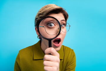 Young woman holding magnifying glass and looking surprised with retro khaki shirt against blue...
