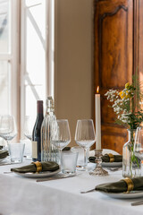 table de diner décorée avec des bougies, une bouteille de vin, verres à pieds, assiettes blanches et serviettes vertes, la lumière entre dans la pièce