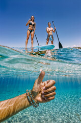 Split shot image taken below the wavy water surface. Thumbs up male hand in bracelet is underwater, two adults are on boards in the foreground