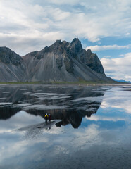A couple adventurer stands amidst the tranquil reflections of Vestrahorn mountain at Stokksnes,...