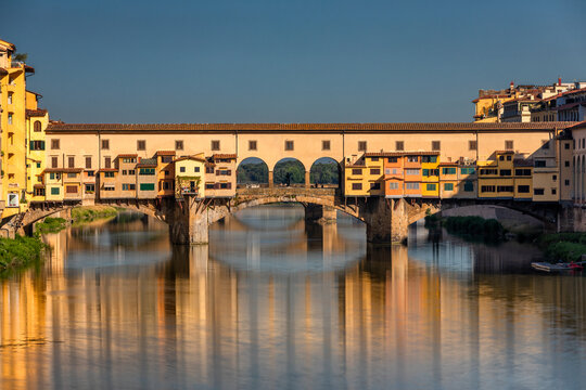 View of the Ponte Vecchio's golden hues reflecting on the tranquil Arno River under a clear sky, creating a timeless scene, Florence, Tuscany, Italy.
