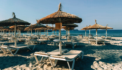 Empty beach with straw parasols and lounge chairs facing the blue sea under clear sky.