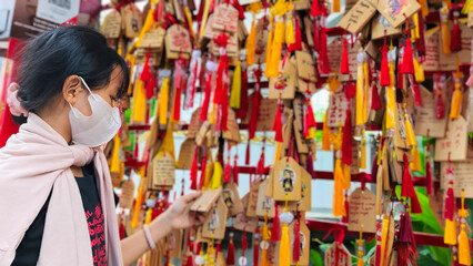 A little girl hangs a wooden sign bearing a wish on a bright red wish wall, adorned with wooden tags and red tassels, a hallmark of Chinese temples and religious sites.