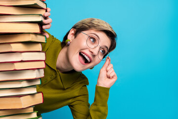Cheerful young woman in stylish casual attire peeks playfully behind stack of book against bright...