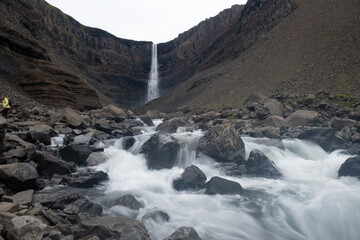 Water flows vigorously over rocks, leading to a stunning Hengifoss waterfall nestled among steep...