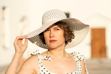 A woman with short curly hair wearing a large white woven hat and a pearl necklace outdoors in...