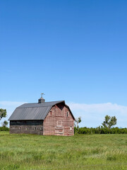 A weathered red barn stands alone in a wide grassy prairie under a vibrant blue sky, evoking...