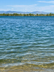 Vibrant blue lake water sparkles under the summer sun with a distant tree-lined shoreline and Montana mountains in the background.
